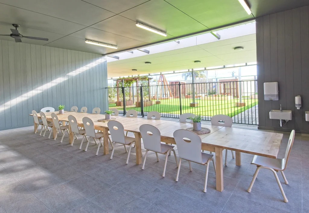 A long wooden table with white chairs is set up in a bright, modern indoor-outdoor space. The area features large windows and a view of an outdoor playground.