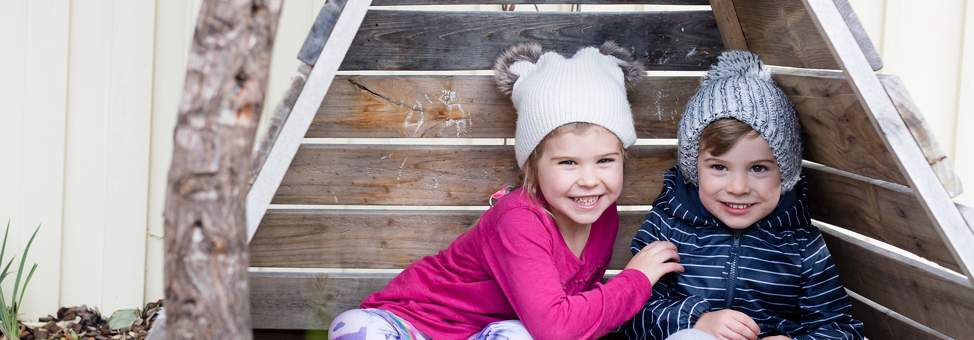 Two young kindergarten children are sitting in a wooden triangular structure, smiling at the camera. The child on the left wears a pink top and a white beanie, while the child on the right has a blue hoodie and grey beanie.