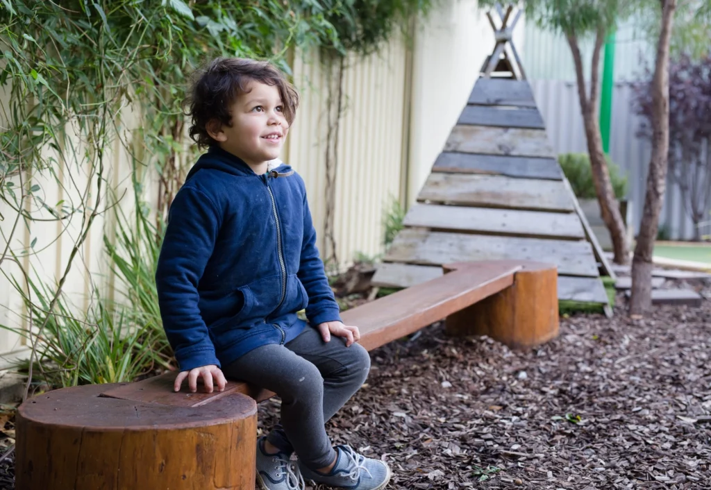 A Kindergarten child in a blue jacket sits on a wooden bench in an outdoor play area with wood chip mulch, greenery, and a wooden teepee structure in the background.