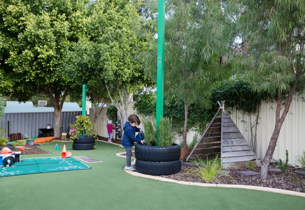 Children play in an outdoor early learning playground with green artificial grass, trees, repurposed tire garden beds, a wooden teepee, and various toys around them.