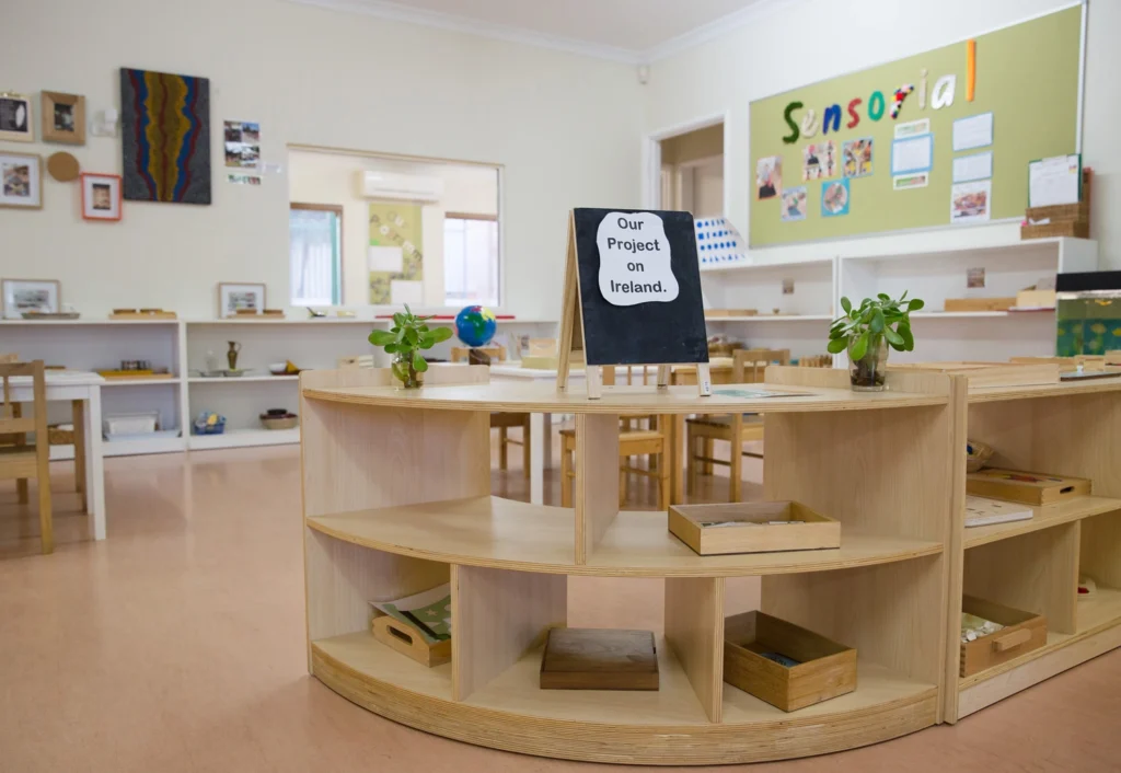 A Stirling childcare classroom with wooden shelves and tables, a small chalkboard says "Our Project on Ireland", and various educational materials and plants neatly arranged.