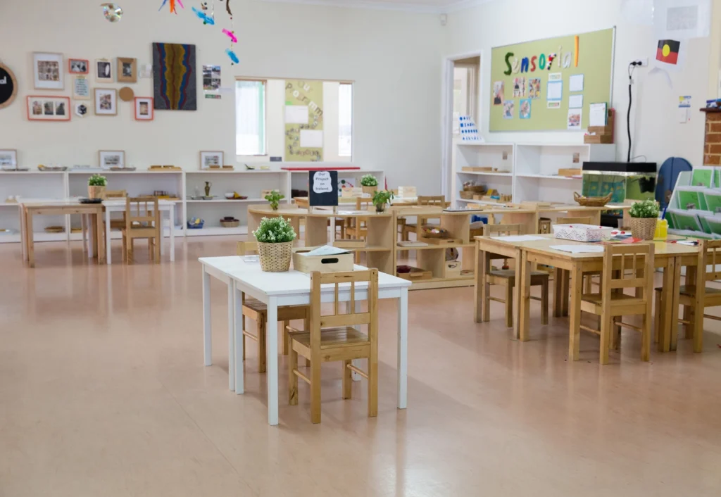 A childcare classroom at Busy Bees Balcatta with small wooden tables and chairs, colorful wall decorations, potted plants, and educational materials neatly arranged on shelves.