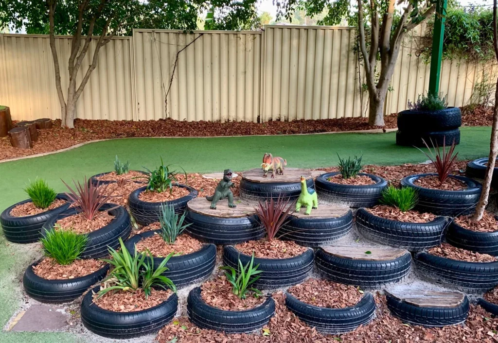 A playground scene at Stirling childcare centre in the suburb of Balcatta. Repurposed tires are arranged in tiers, filled with mulch, various plants and toy dinosaurs.