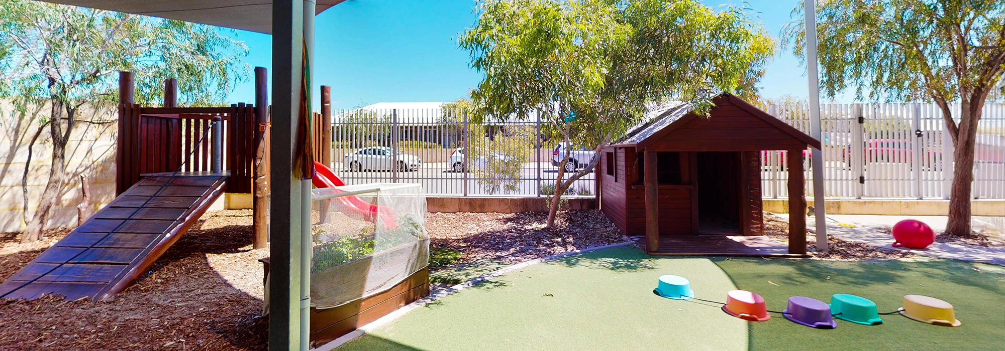 Outdoor playground area with a wooden play structure, small house, slides, climbing ramp, and colorful stepping stones on artificial grass and mulch. Trees provide shade in the background.