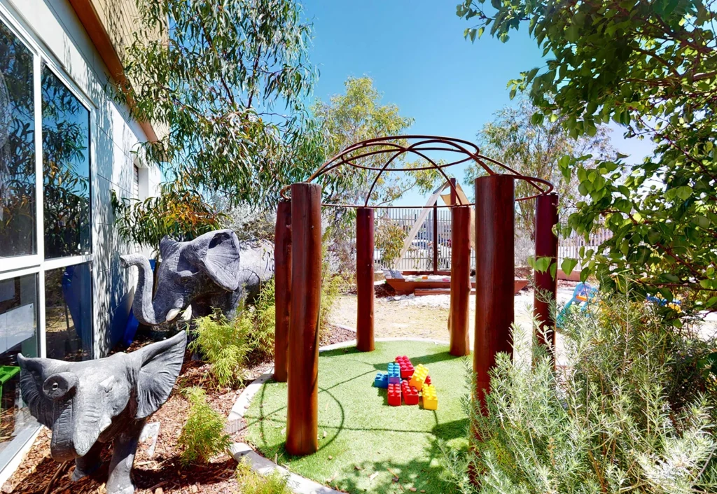 Outdoor play area with an elephant statue, greenery, and a grassy section with large toy blocks and a metal framework structure.