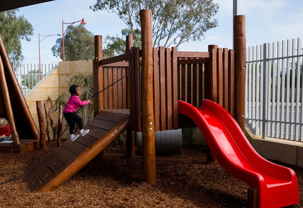 A child in a pink jacket climbs a wooden ramp of a playground structure with a red slide nearby. Trees and a metal fence are in the background.