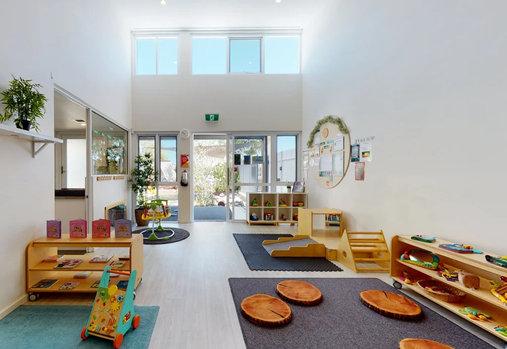 A bright, spacious room with large windows, various children's play furniture, bookshelves, toys, and plants. Mats and cushions are placed on the floor, creating a welcoming environment for children.