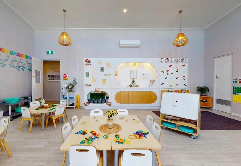 A clean, well-lit classroom with tables and chairs, educational toys, wall decorations, and colorful art supplies neatly arranged for children's activities.