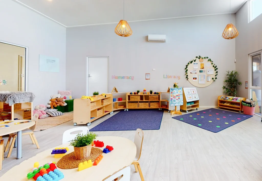 A well-organized, brightly lit classroom with children's toys, bookshelves, and colorful play mats. Wooden furniture and decorations create a warm, inviting atmosphere.