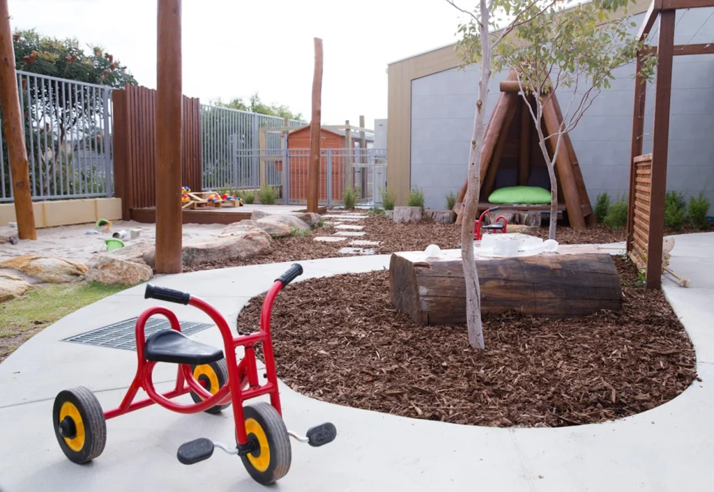 A playground at the day care featuring a red tricycle, wooden climbing structures, mulch ground cover, and a tree.