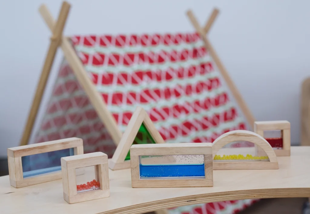 Wooden blocks with beads inside are displayed on a table, perfect for early learning activities. In the background, there is a small tent with a red and white patterned fabric cover, creating an inviting atmosphere for any kindergarten or childcare setting.