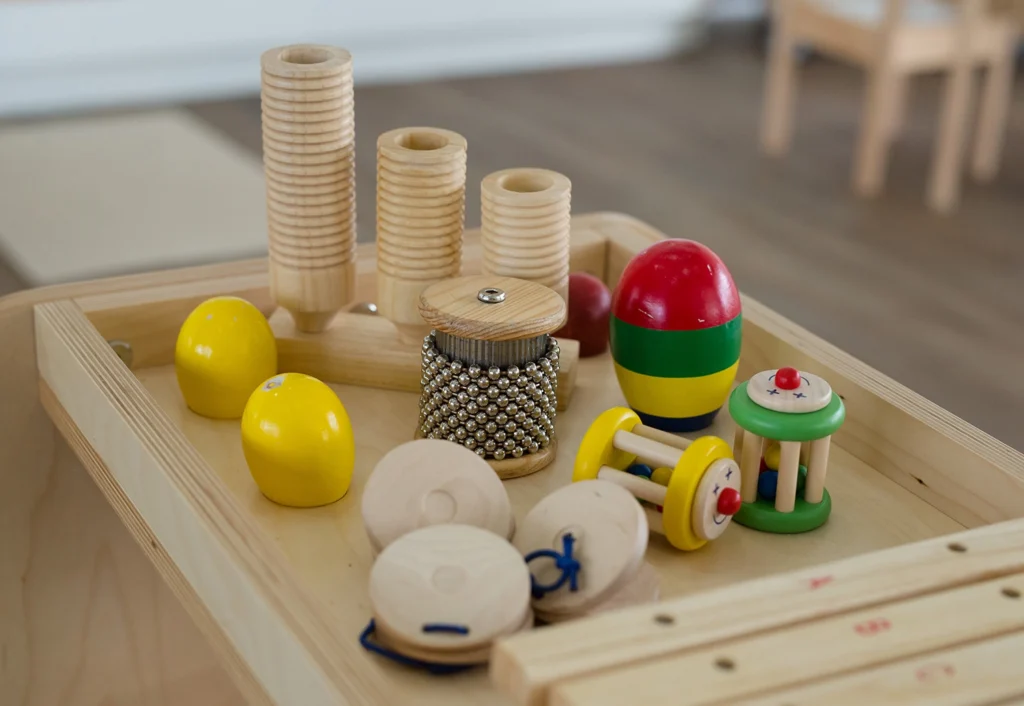 A collection of various wooden and plastic toys, including stacking rings, maracas, and a rattle, ideal for early learning in childcare or preschool settings, are placed on a wooden table.