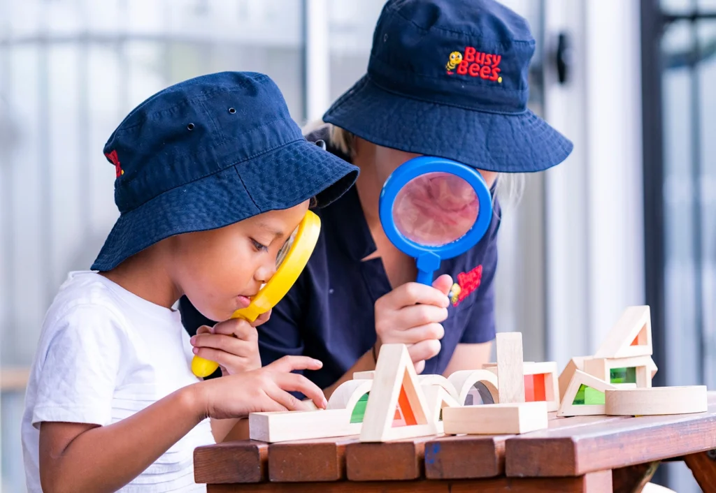 A child and Early Childhood Teacher children wearing navy bucket hats explore wooden blocks with magnifying glasses at a table, immersing themselves in early learning activities.