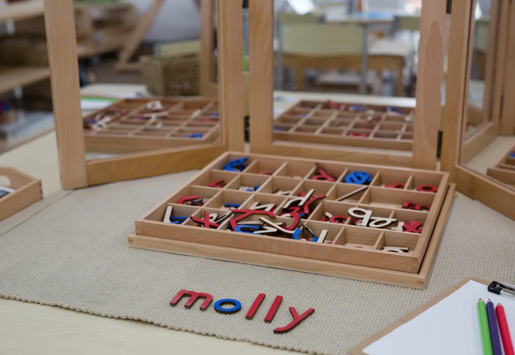 A wooden tray with assorted colorful letters on a table, including letters spelling "molly" in red. Perfect for early learning and kindergarten, the setup also features mirrors around and colored pencils nearby, creating an engaging environment for childcare activities.