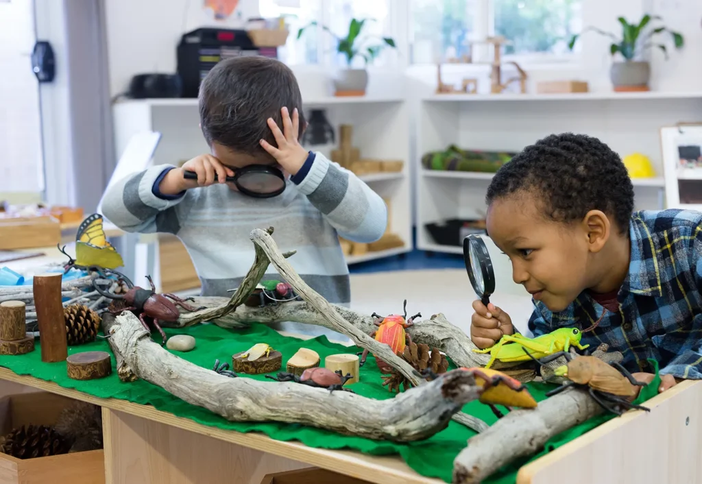 Two young children use magnifying glasses to explore a nature-themed sensory table with logs, toy insects, and natural materials in a preschool classroom setting.