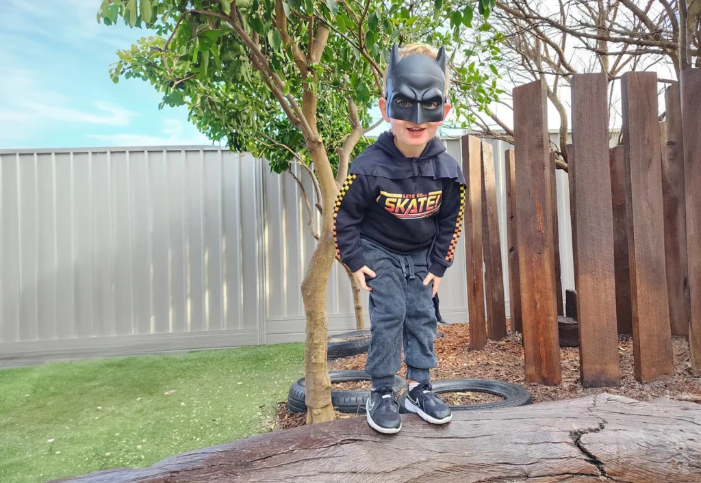 A child dressed in a Batman mask stands on a wooden log in the outdoor playground at Princeton Pre School in Osborne Park.