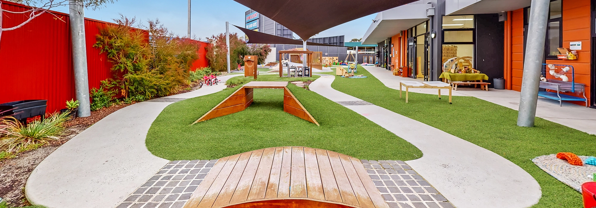 A modern daycare playground featuring artificial turf, wooden play structures, shaded areas, and various children's toys. Pathways and greenery surround the outdoor space.