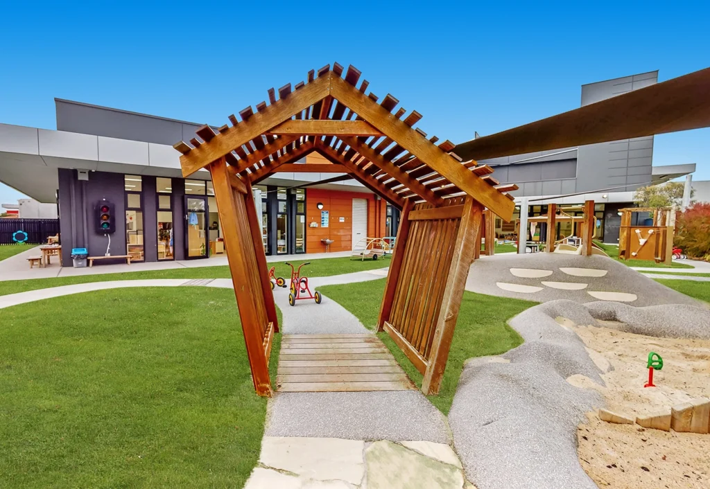 Wooden structure in a modern playground with green grass, sandpit, tricycles, and a building in the background under a clear blue sky.