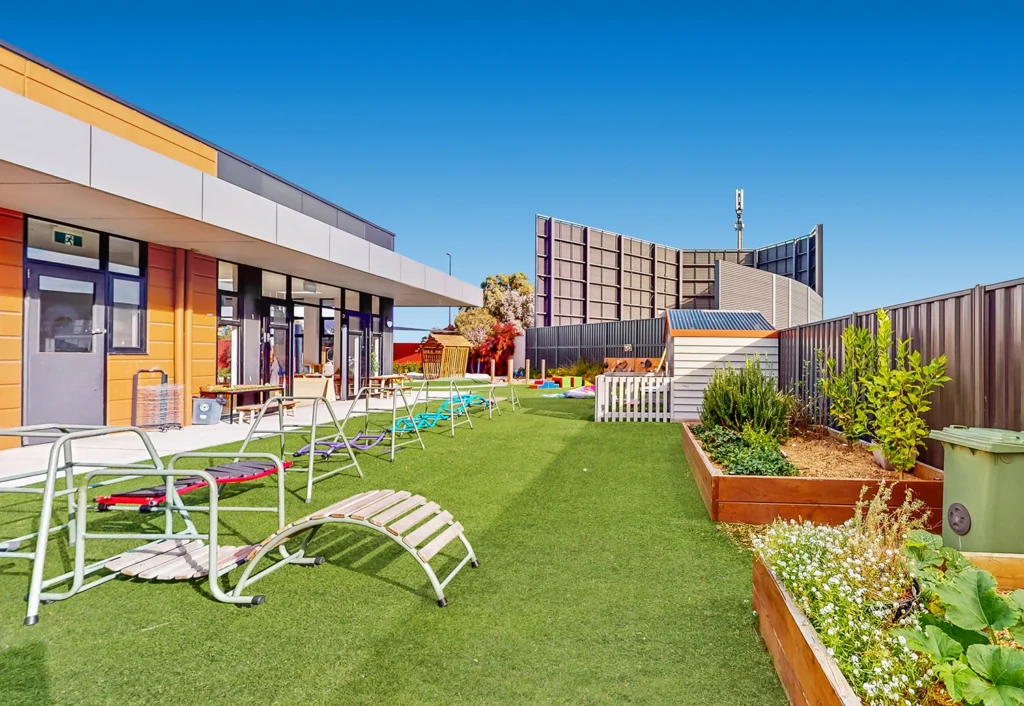 Outdoor area of a childcare center with artificial turf, playground equipment, raised garden beds, and seating. Modern building structures and a clear blue sky in the background.