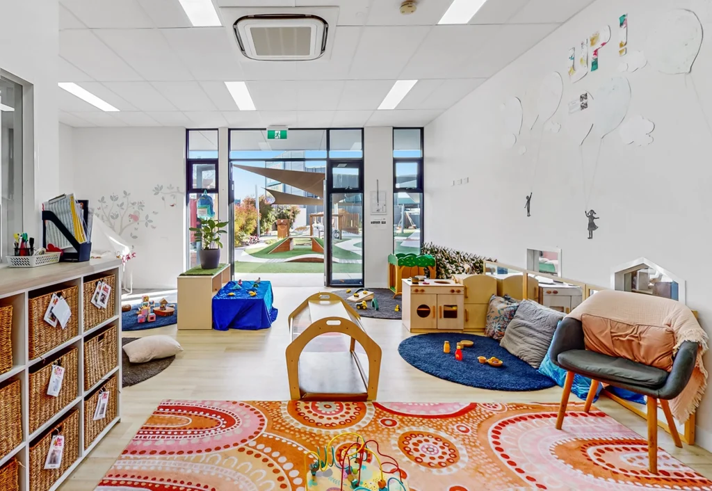 A brightly lit classroom with a colorful rug, wooden furniture, toys, and play areas. Large windows provide a view of an outdoor play area.