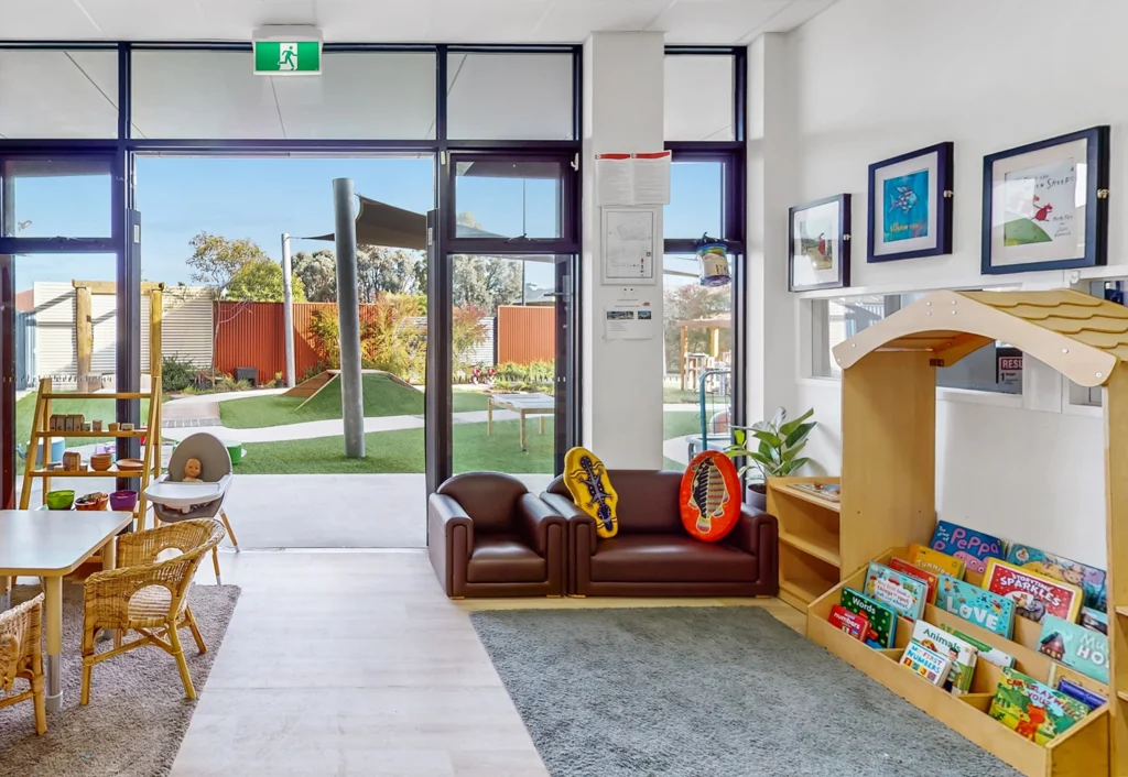 A bright children's reading and play area with small furniture, books, artwork on the walls, and a view of an outdoor playground through large windows.