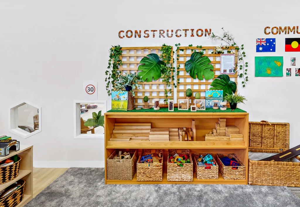 Classroom construction area with wooden blocks, baskets of building materials, and greenery décor. A display board features plants, flags, and children's art. Storage baskets are neatly arranged below.