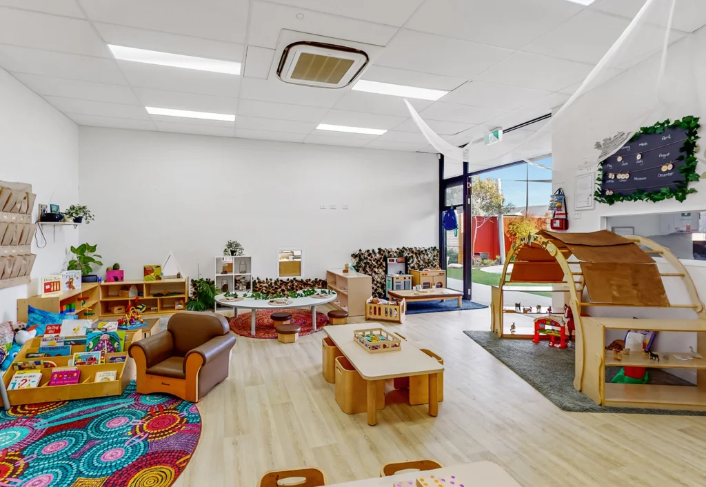 A brightly lit preschool classroom with various play areas, including a reading nook, activity tables, and a circular rug. Shelves with books and toys line the walls. Large windows provide ample natural light.