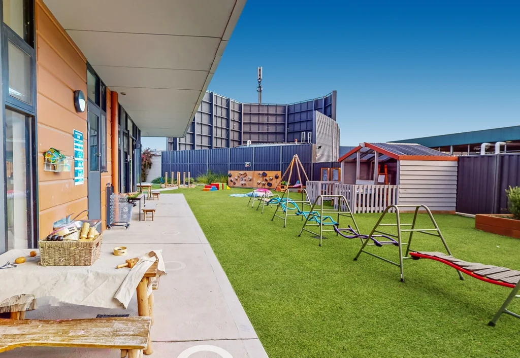 An outdoor playground at a daycare center with slides, swings, and a playhouse on artificial grass, adjacent to a building.