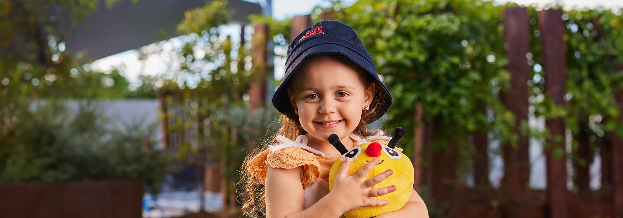 A young girl in a navy blue hat smiles and holds a yellow stuffed toy outdoors, perhaps during a fun day at kindergarten. The background includes lush greenery and a partly cloudy sky.