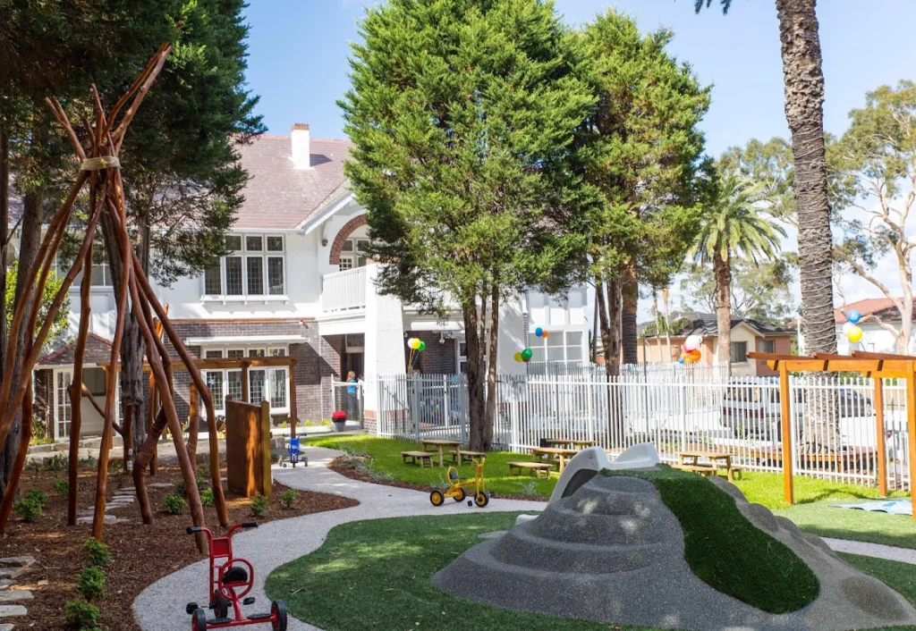 Outdoor playground featuring a teepee, tricycles, a small hill with artificial grass, a walking path, picnic tables, and balloons tied to a white fence. A large house is visible in the background.