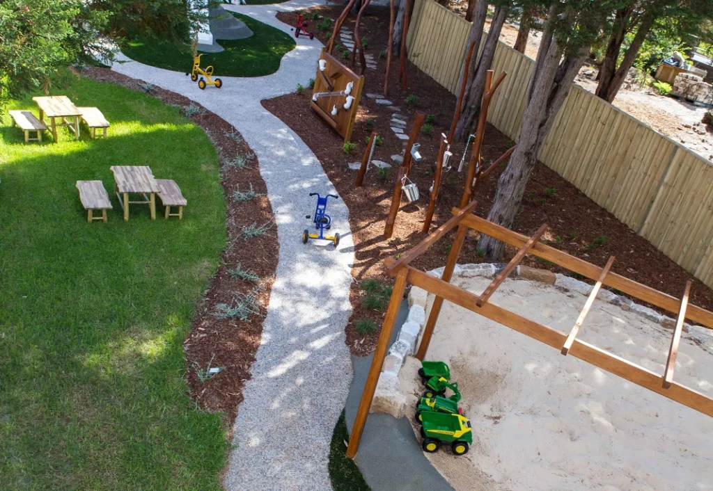 Aerial view of a children's playground featuring a sandbox, toy vehicles, picnic tables, and various play structures. The area is surrounded by trees and lined with a wooden fence.