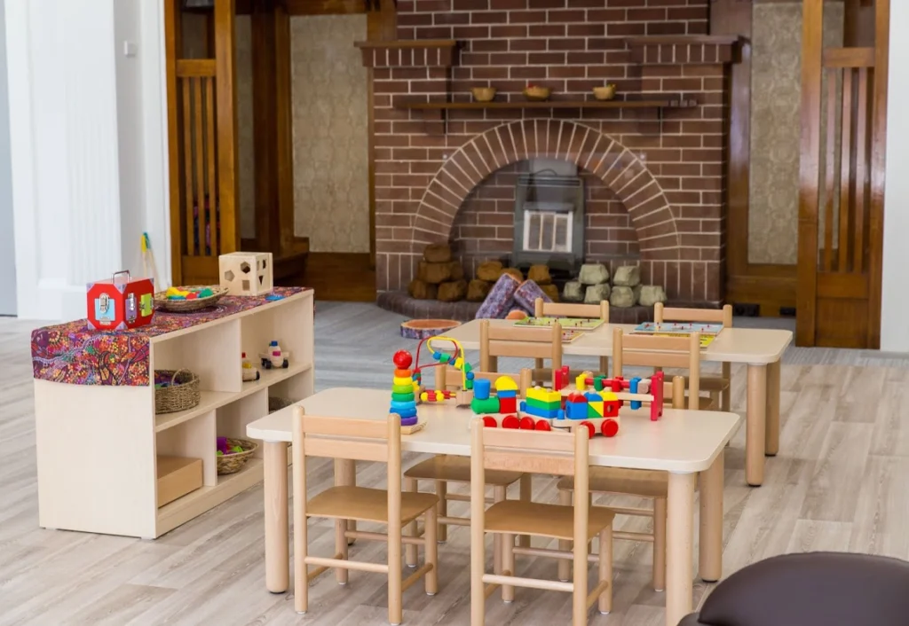 A playroom with a brick fireplace, wooden shelves, small tables with colorful toys, and tiny wooden chairs set on a light-colored floor.