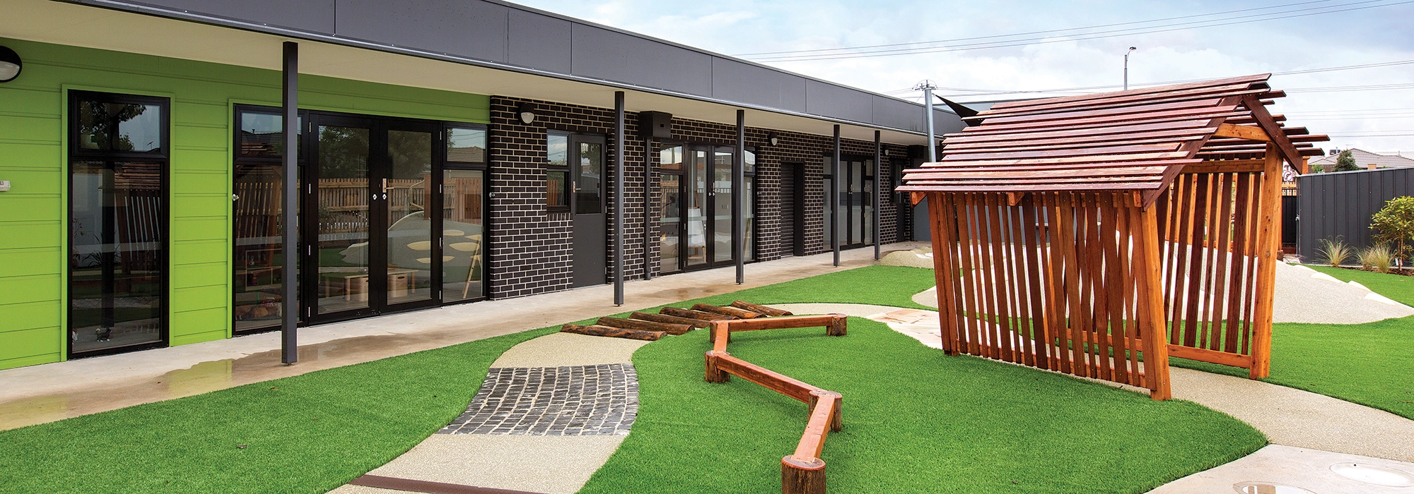 A playground area featuring a wooden play structure, artificial turf, and a path of stepping stones, adjacent to a row of modern building units with glass doors and windows—ideal for inspiring early learning and exploration among preschool children.