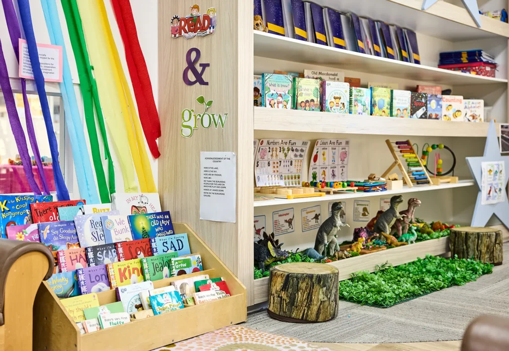 Children's reading area with colorful shelves filled with books, toy dinosaurs, and educational materials. A rainbow curtain decorates one side, and a sign reads "READ & grow. Perfect for preschool or kindergarten settings.