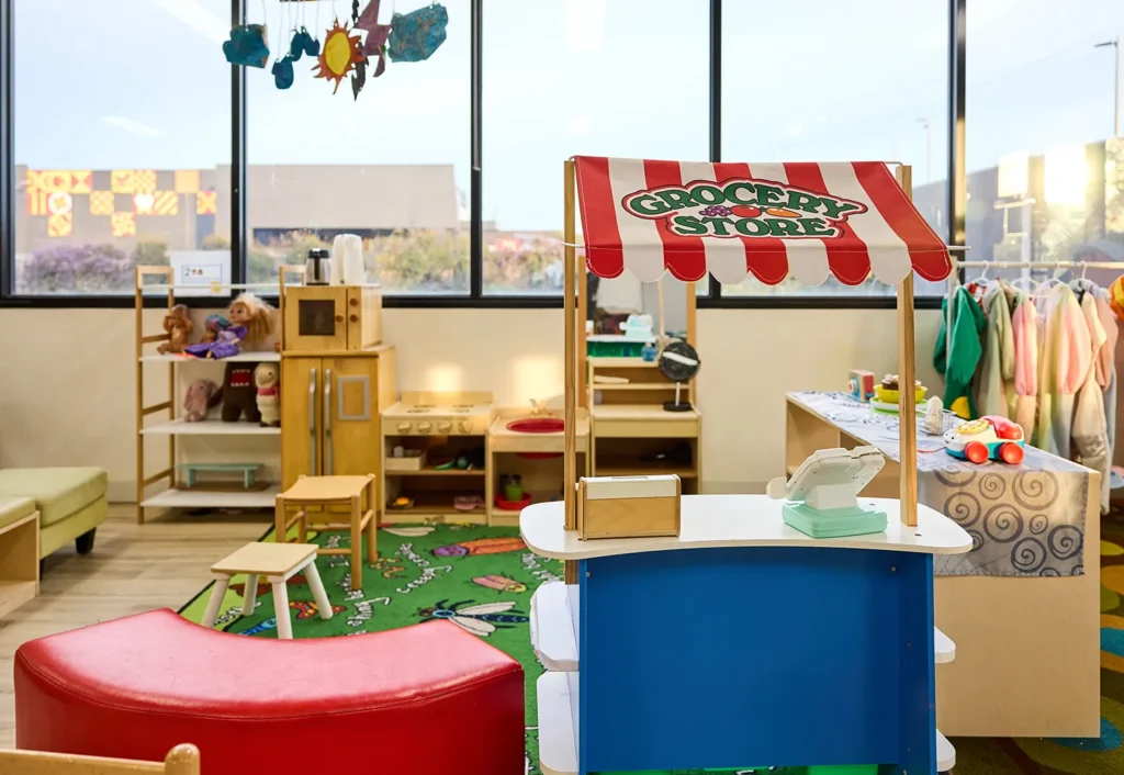 A children's play area featuring a toy grocery store stand, shelves with toys, a rack of children's clothing, and colorful decor ensures an engaging early learning environment. Large windows allow natural light to brighten the space, perfect for any childcare or kindergarten setting.