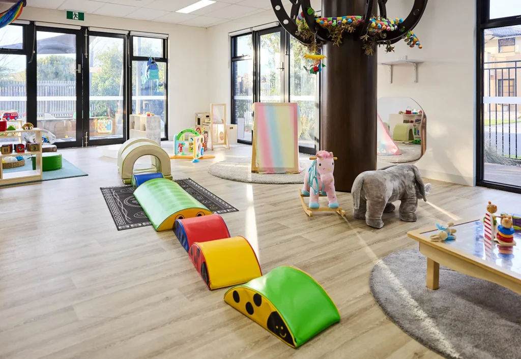 A brightly lit daycare room offers exceptional childcare with play equipment, including a soft climbing structure, a plush unicorn, a plush elephant, and several educational toys on a low table. Large windows allow natural light to fill this inviting preschool space.