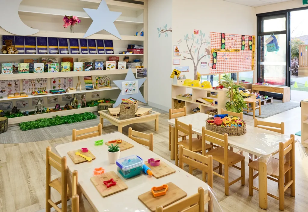 A well-organized early learning classroom with small wooden tables and chairs, shelves filled with books, toys, and learning materials, and vibrant wall decorations.