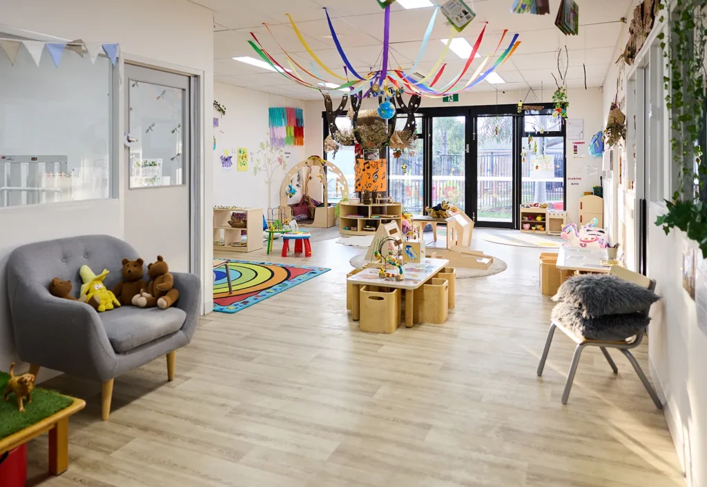 A brightly lit, colorful kindergarten classroom with various play areas, toys, and crafts. A gray sofa with stuffed animals is on the left, and a large window provides ample natural light, creating an inviting space for early learning.