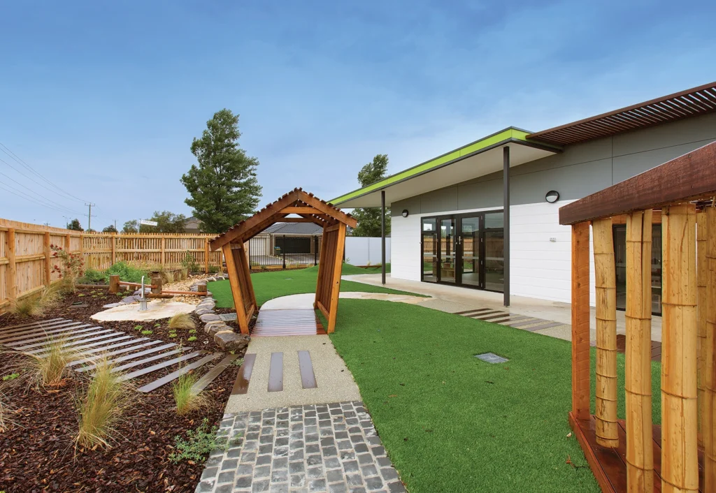 A modern outdoor area at the preschool features a small wooden bridge, a walkway, landscaping with plants and rocks, and a building with large glass doors and a green roof trim in the background.