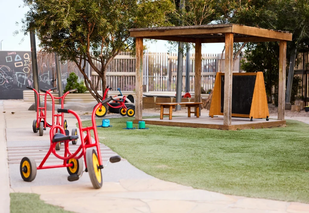 An outdoor play area for early learning features red tricycles, a chalkboard, scattered buckets, and a wooden pavilion with benches, all set on artificial grass and concrete.