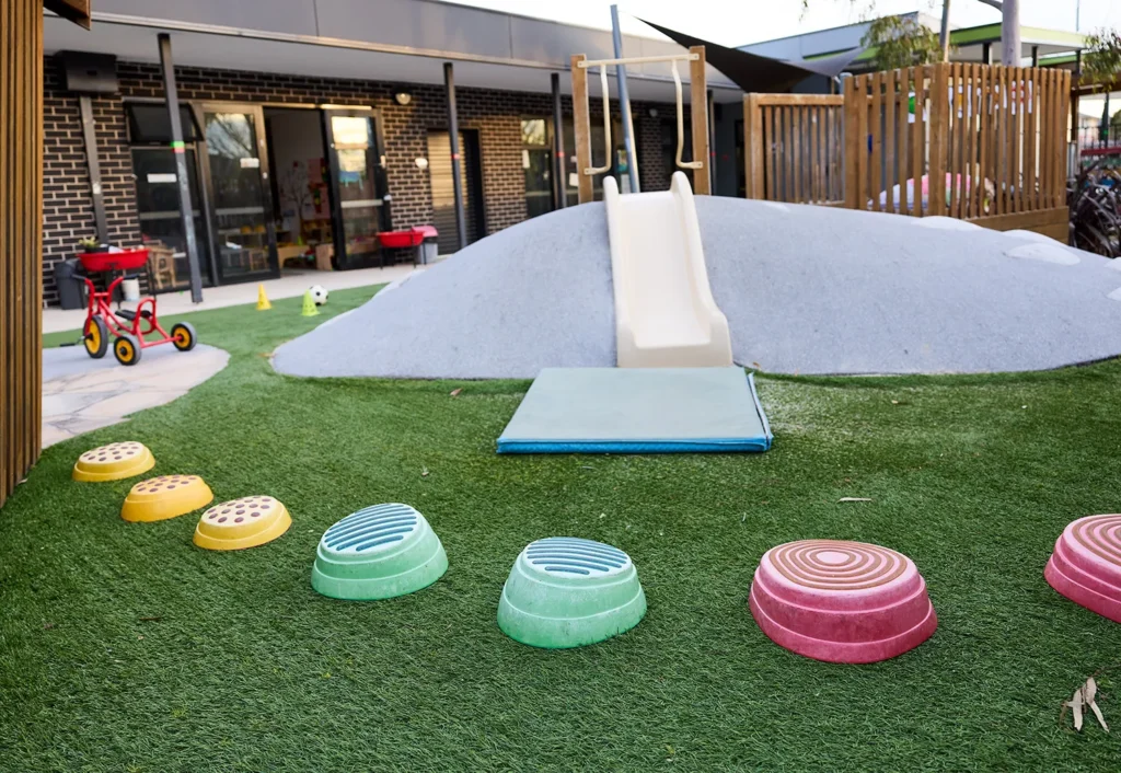 A vibrant playground with a slide, artificial grass, colorful stepping pods, and a tricycle sits invitingly in front of a brick building dedicated to early learning.