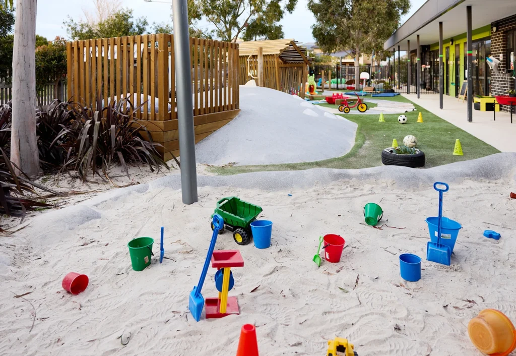 A playground area with a sandbox filled with various colorful toys, including buckets, shovels, and toy trucks. In the background of this early learning space at the day care, there is a grassy section dotted with small ride-on vehicles and structures.