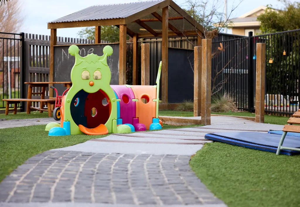 A brightly colored caterpillar-shaped play tunnel in an outdoor playground area with artificial grass, a paved path, and a small wooden shelter in the background, perfect for preschool early learning activities.