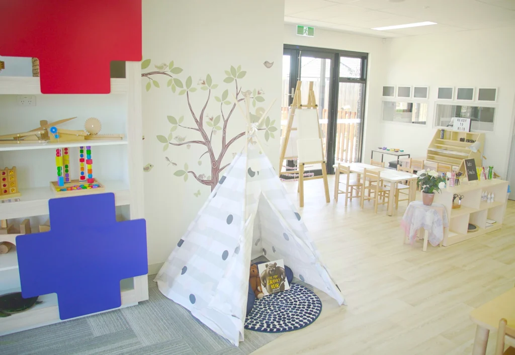 A brightly lit classroom designed for early learning features a small tent, art easels, tables, chairs, storage shelves with toys, and a tree mural on the wall. This vibrant space embodies the essence of quality childcare.