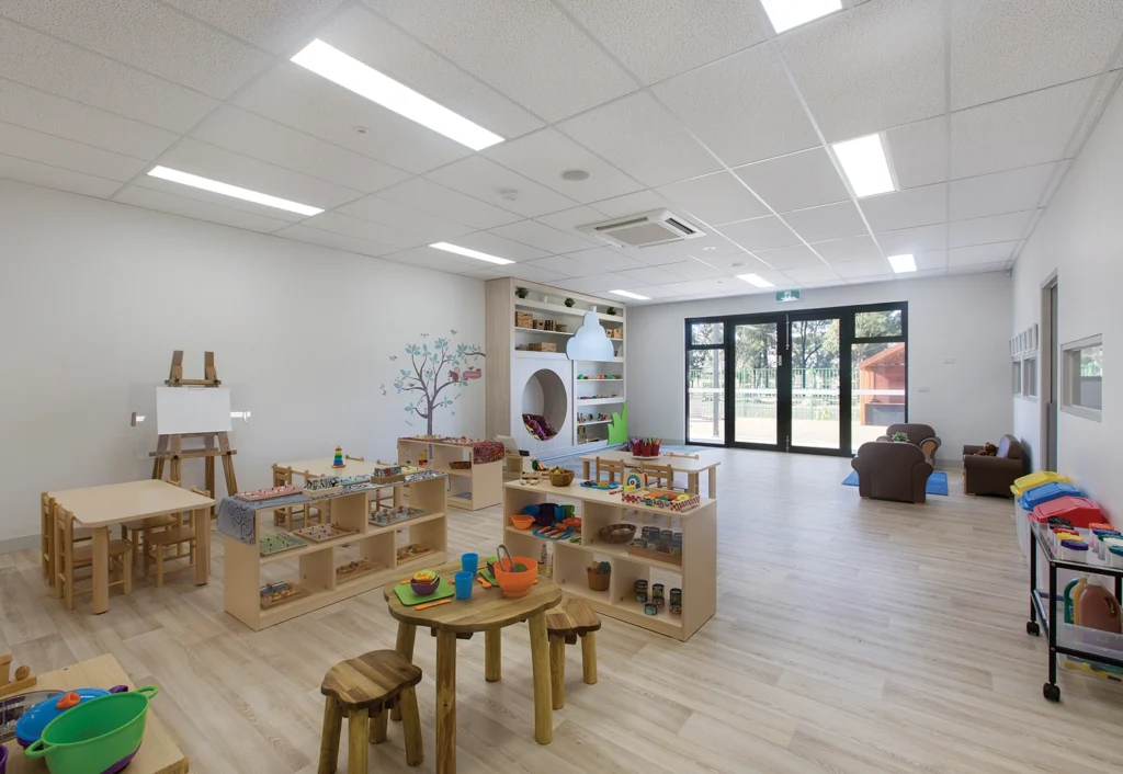 A spacious, well-lit daycare classroom with various learning stations, including art supplies, building blocks, and a reading nook, is arranged across wooden tables and shelves on a light wood floor. Perfect for fostering early learning in a nurturing environment.