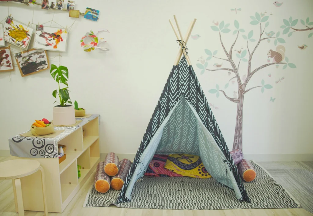 A small teepee tent with patterned fabric is set up in a children's playroom, perfect for early learning and preschool activities. It stands alongside a bookshelf with toys, a green plant, and a wall decorated with a mural tree and children's art.