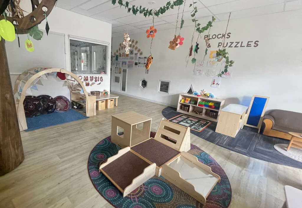 A child-friendly, preschool room with labeled sections for blocks, puzzles, and music. The day care area is furnished with play mats, shelves, wall decorations, and various play structures.