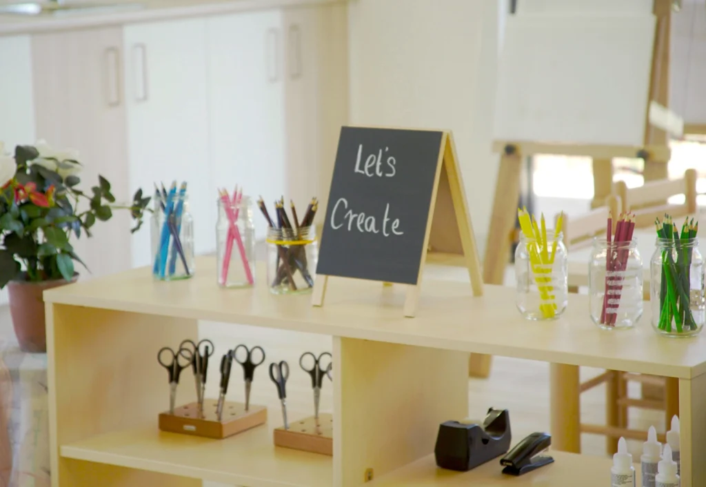 A table with craft supplies including scissors, colored pencils, and a small blackboard sign that reads "Let's Create" in a preschool or childcare classroom setting.