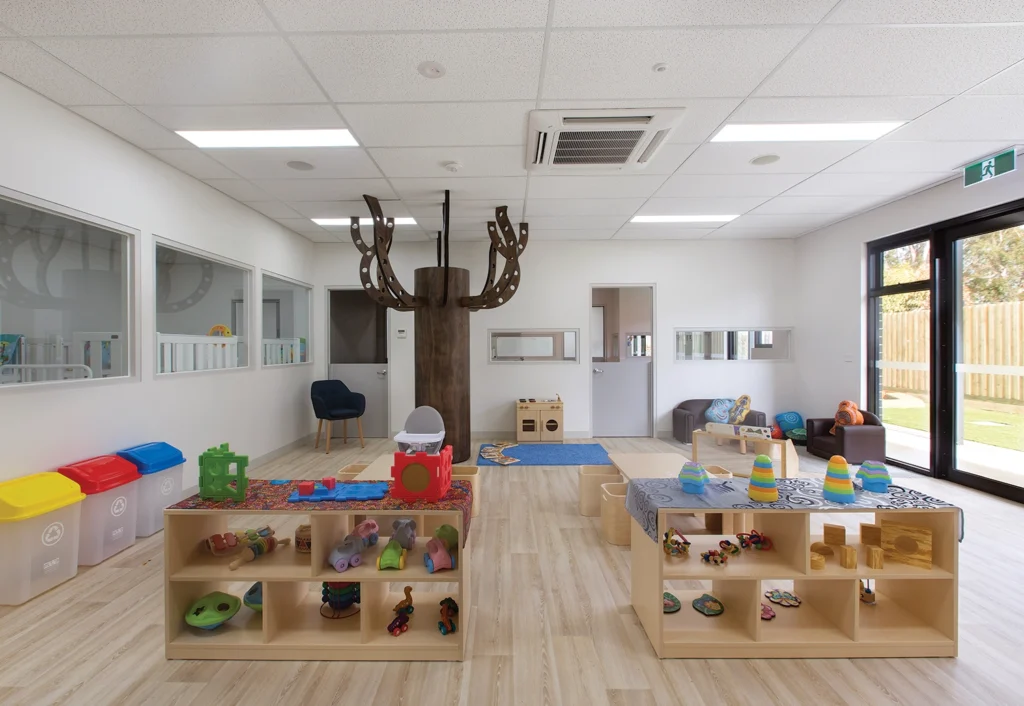 A spacious, modern daycare room designed for early learning, featuring wooden shelves holding various toys, a tree-like indoor play structure, colorful bins for recycling, and large windows allowing natural light.