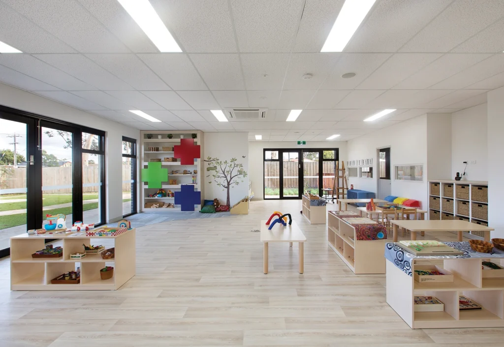 A spacious, well-lit preschool classroom features wooden furniture, educational toys, colorful wall decorations, and large windows overlooking a green outdoor area.